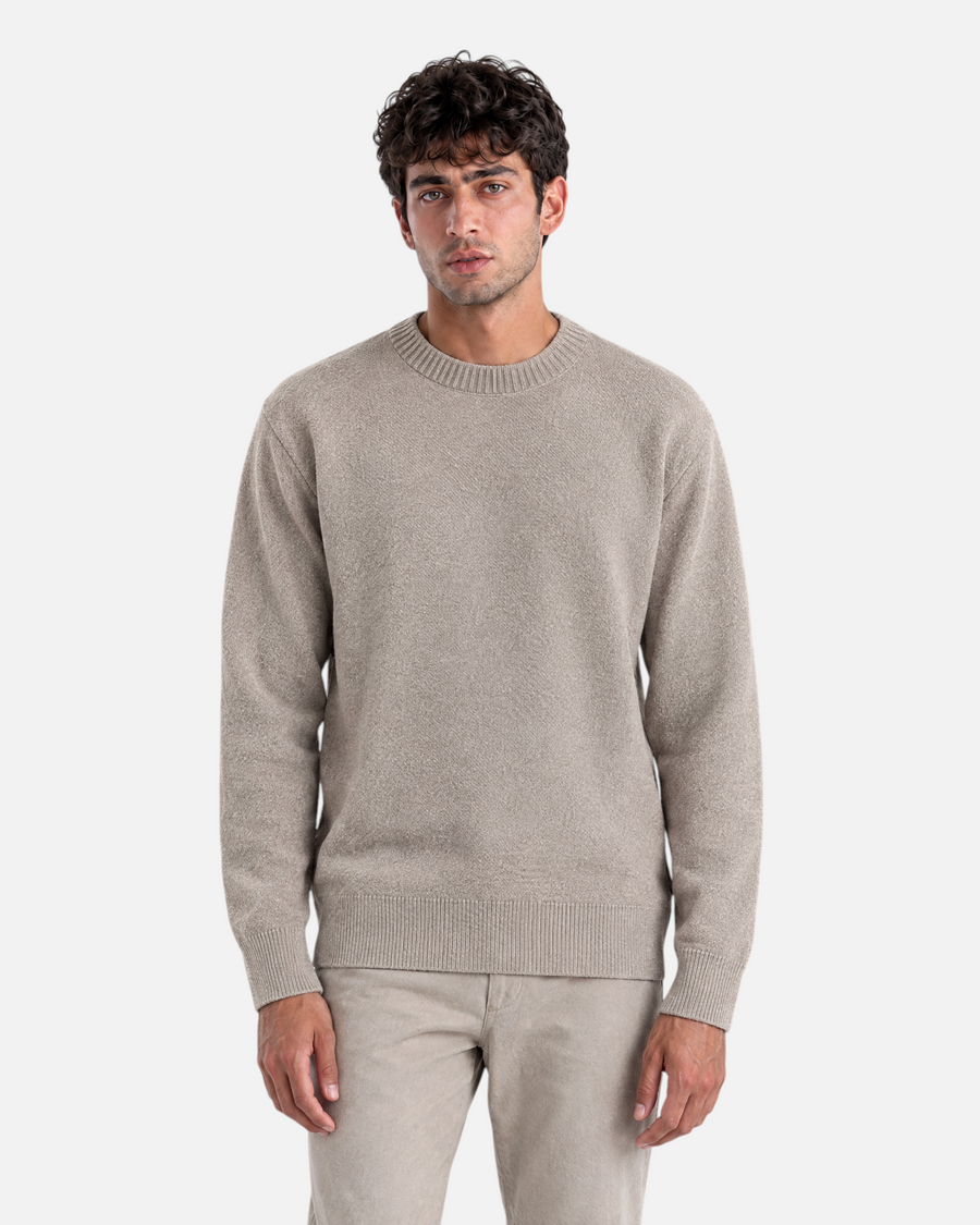 A man wearing a beige RUVR knit pullover with a ribbed crew neck, cuffs, and hem against a grey background.